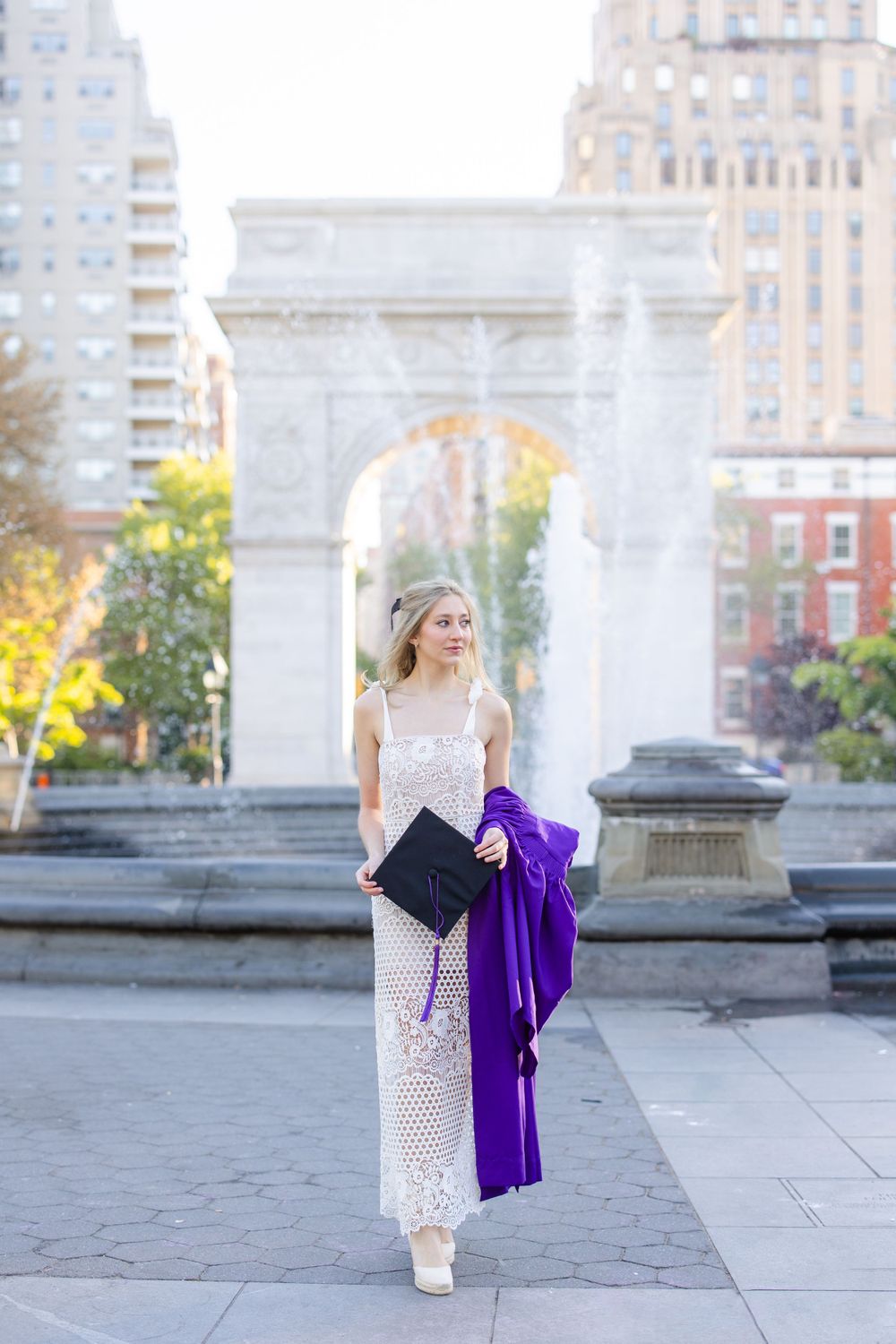 Graduate poses with diploma and purple gown by Washington Square Park arch.