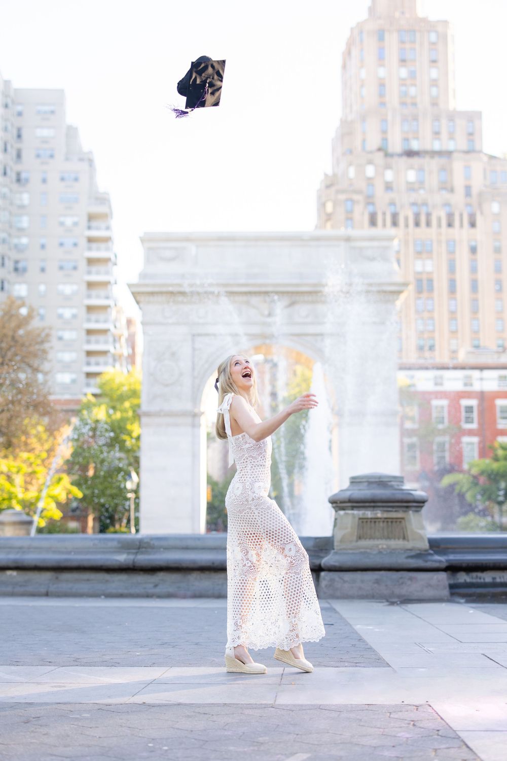 Graduate in a white lace dress tosses cap in front of Washington Square Arch in NYC.