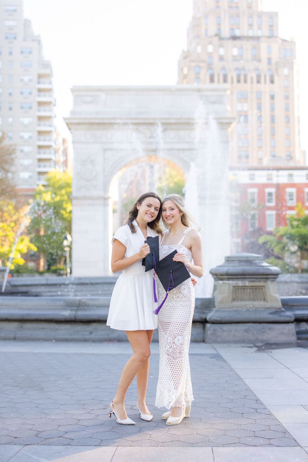 Two graduates pose together in white dresses at Washington Square Park.