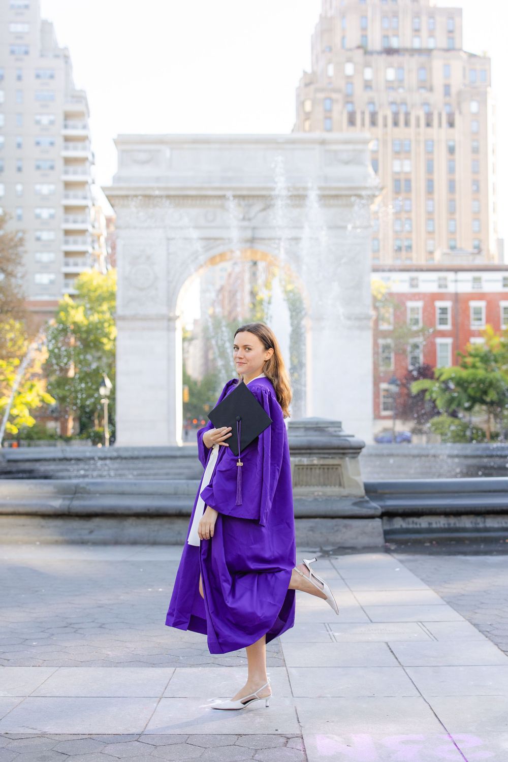 Graduate in purple NYU graduation gown poses in front of Washington Square Arch.
