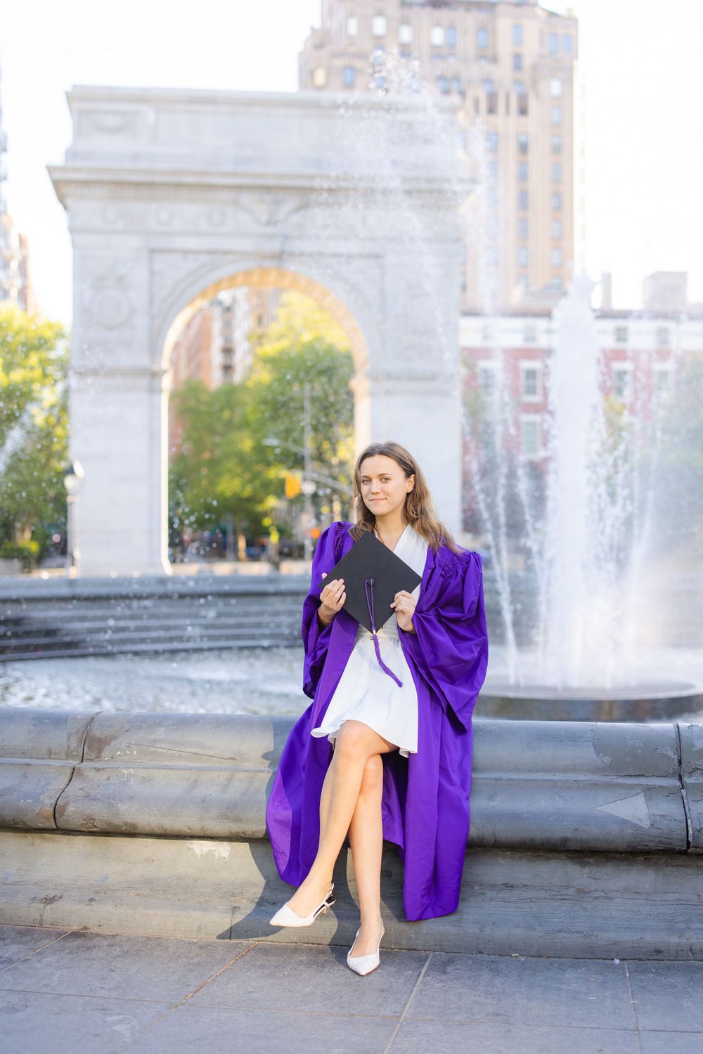 Graduate in purple commencement robe sits on fountain edge at Washington Square Park.