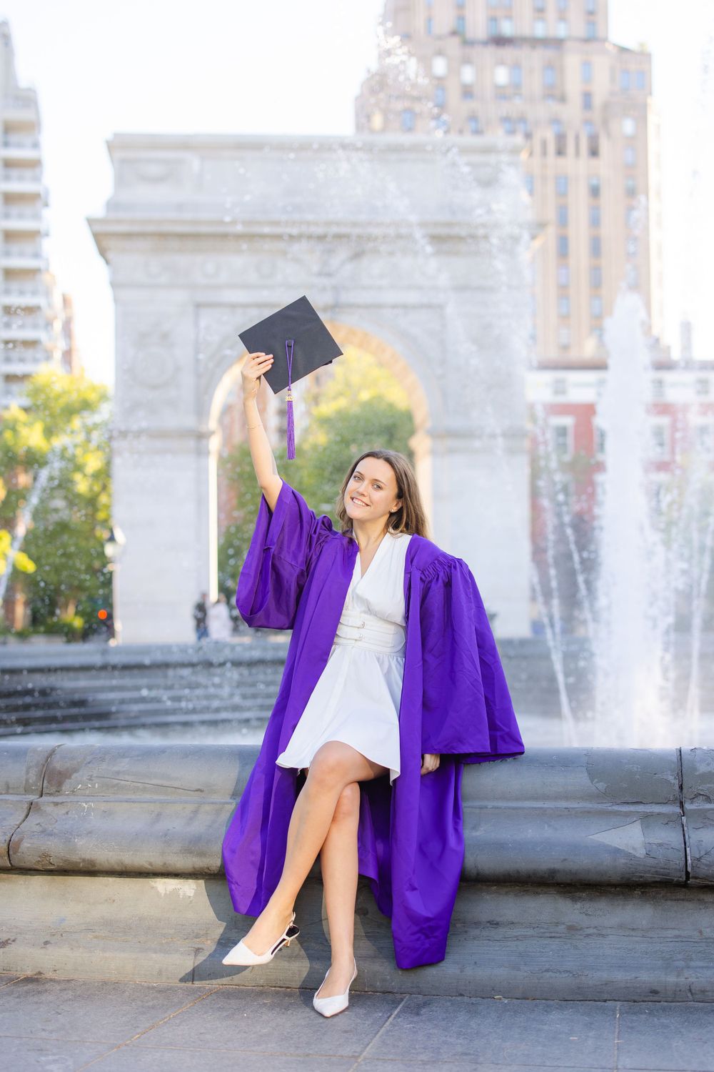 Graduate in purple gown holds up diploma while posing by Washington Square fountain.