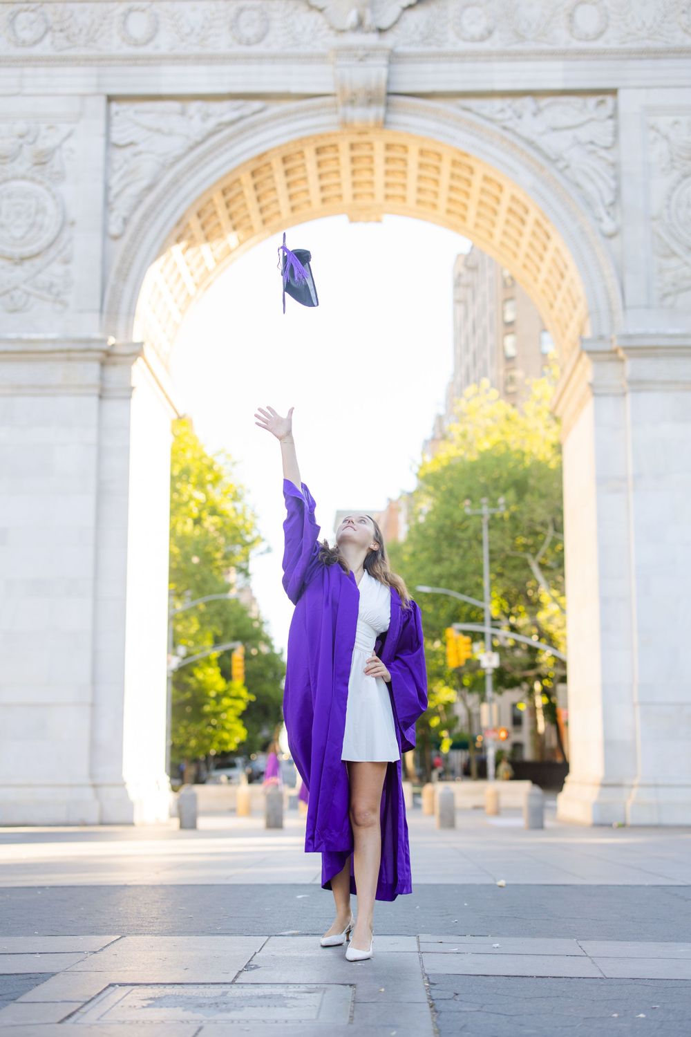 Graduate throws cap in the air under Washington Square Arch in purple graduation robes.