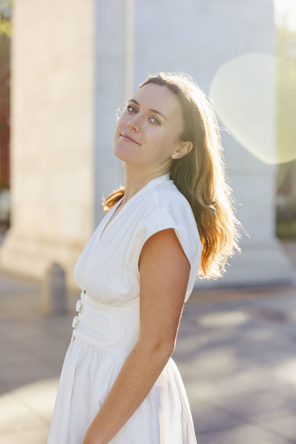 Woman in white dress poses for graduation portrait in sunlight.