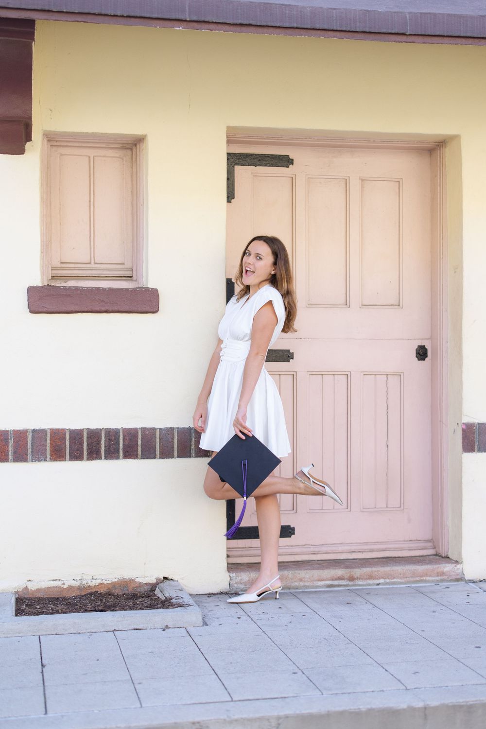 Graduate in white dress poses with diploma by pink door.