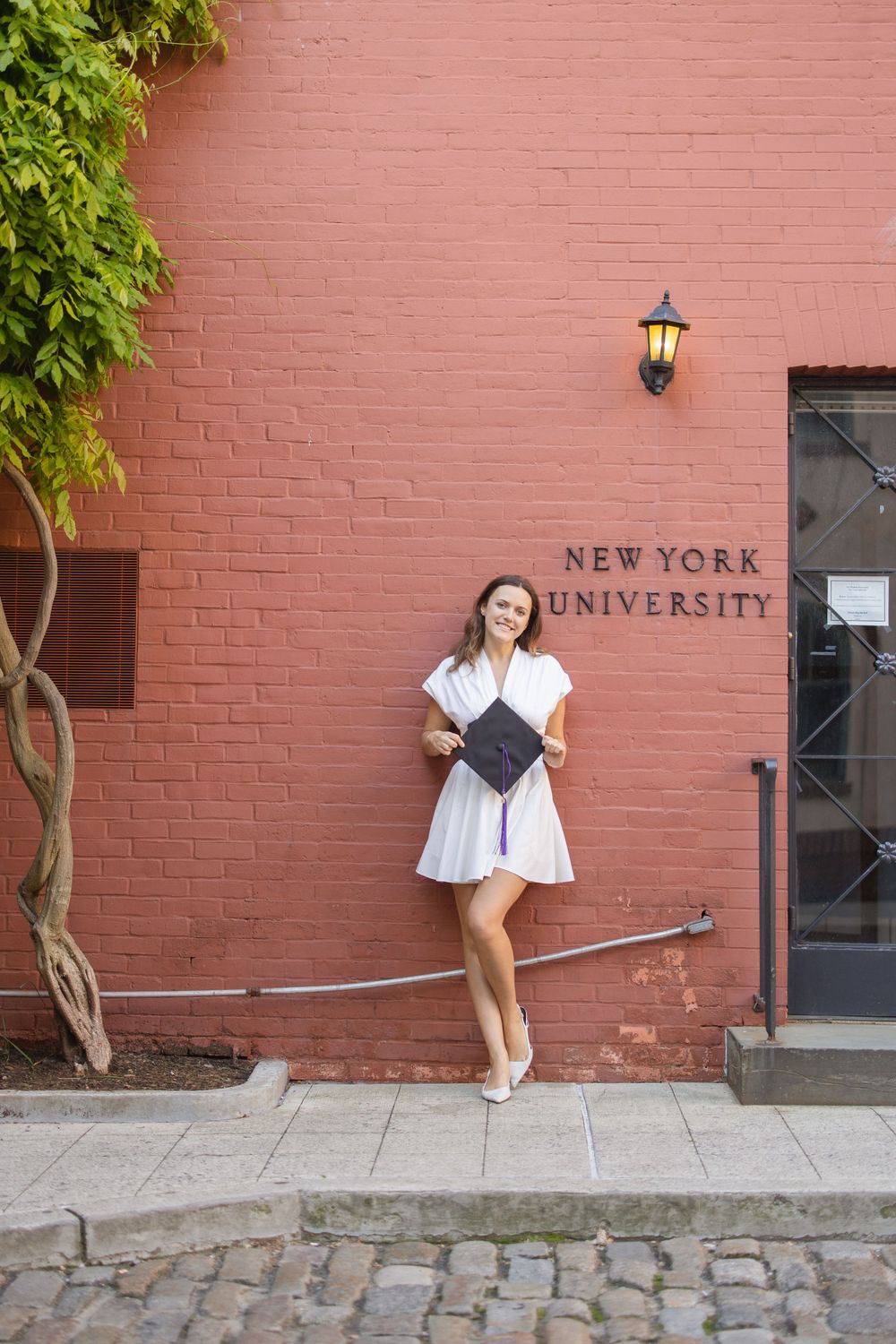 Graduate posing in a white dress against a pink brick wall with New York University signage.