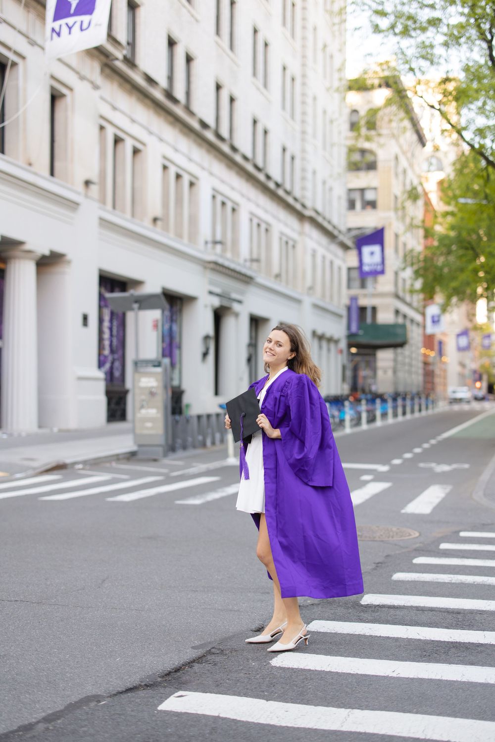 Graduate in purple gown poses on NYU campus street.
