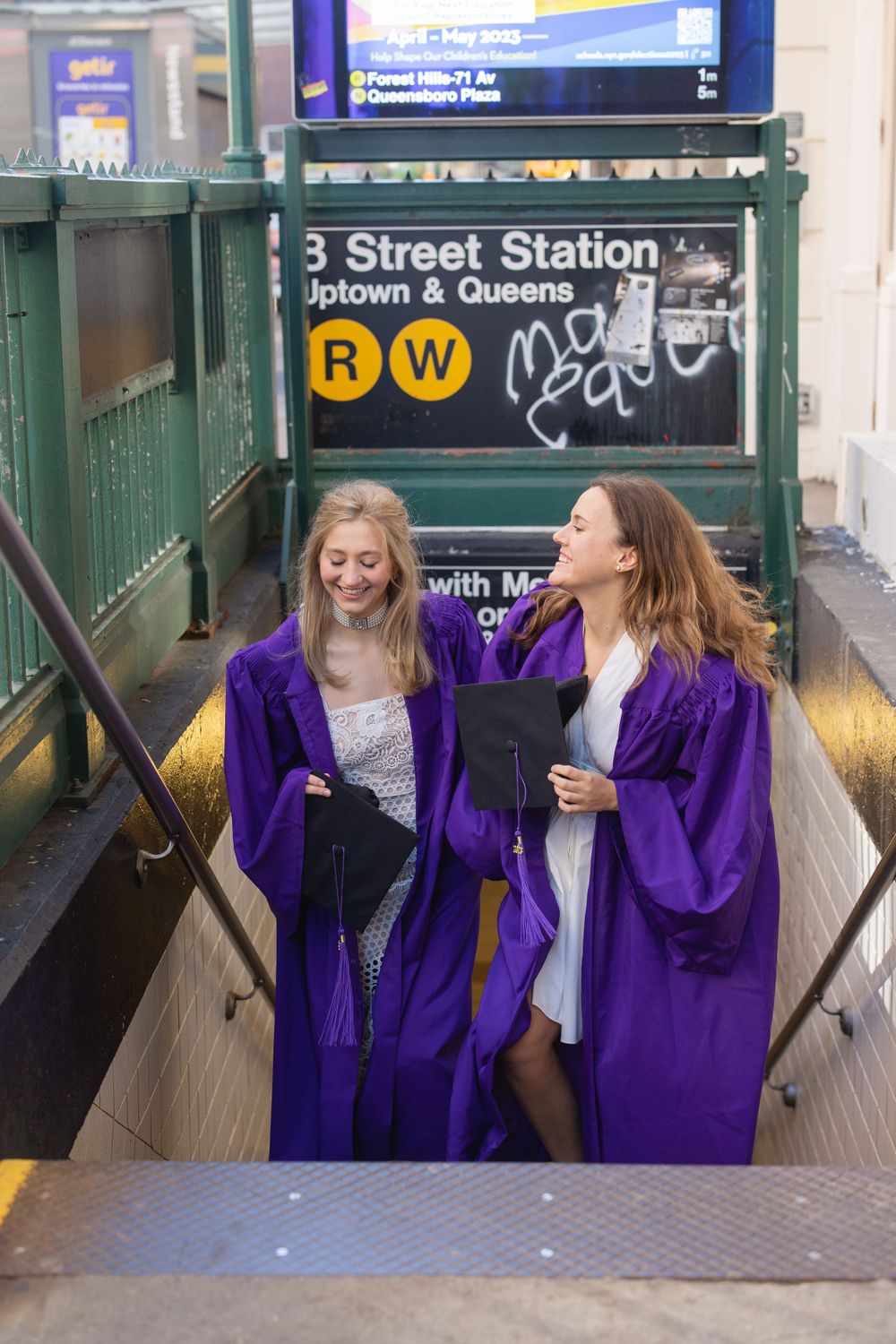 Two graduates in purple robes celebrating at a subway station entrance.