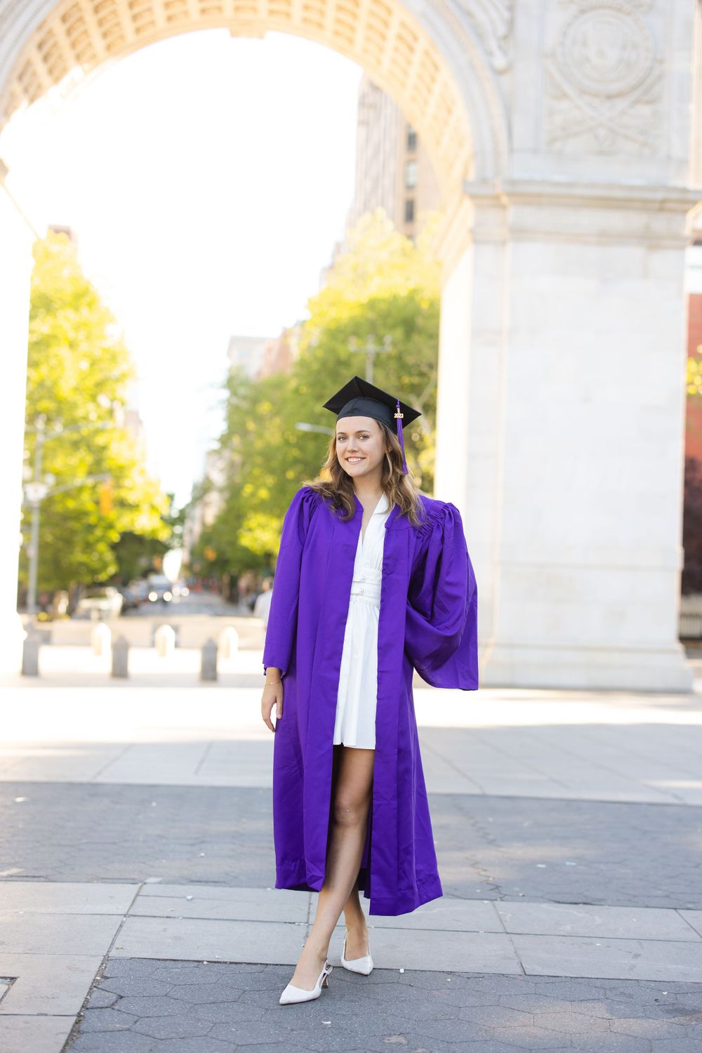 Graduate in purple graduation regalia posing under Washington Square Arch on a sunny day.