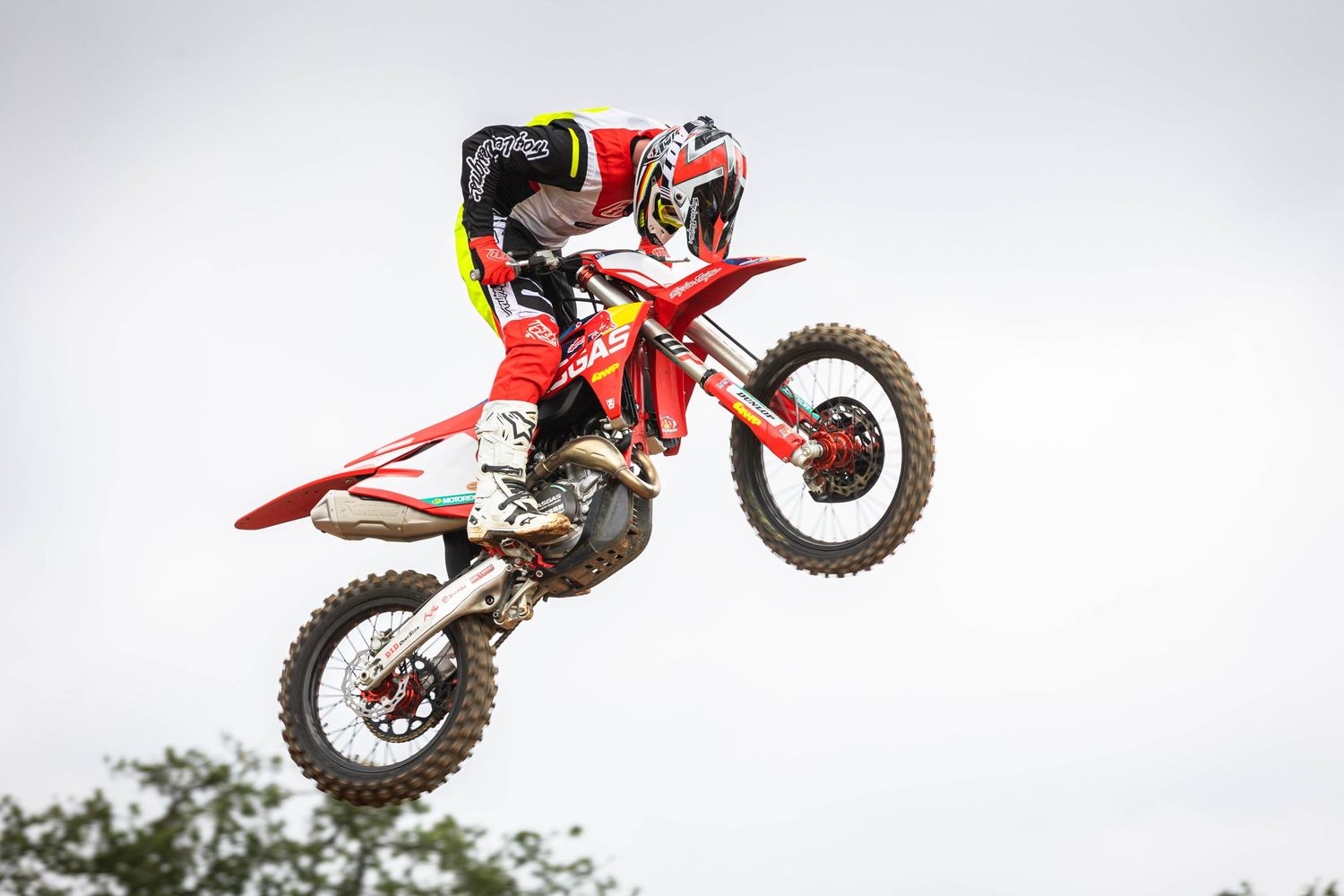 Motorcross rider performs aerial stunt on red dirt bike against cloudy sky.
