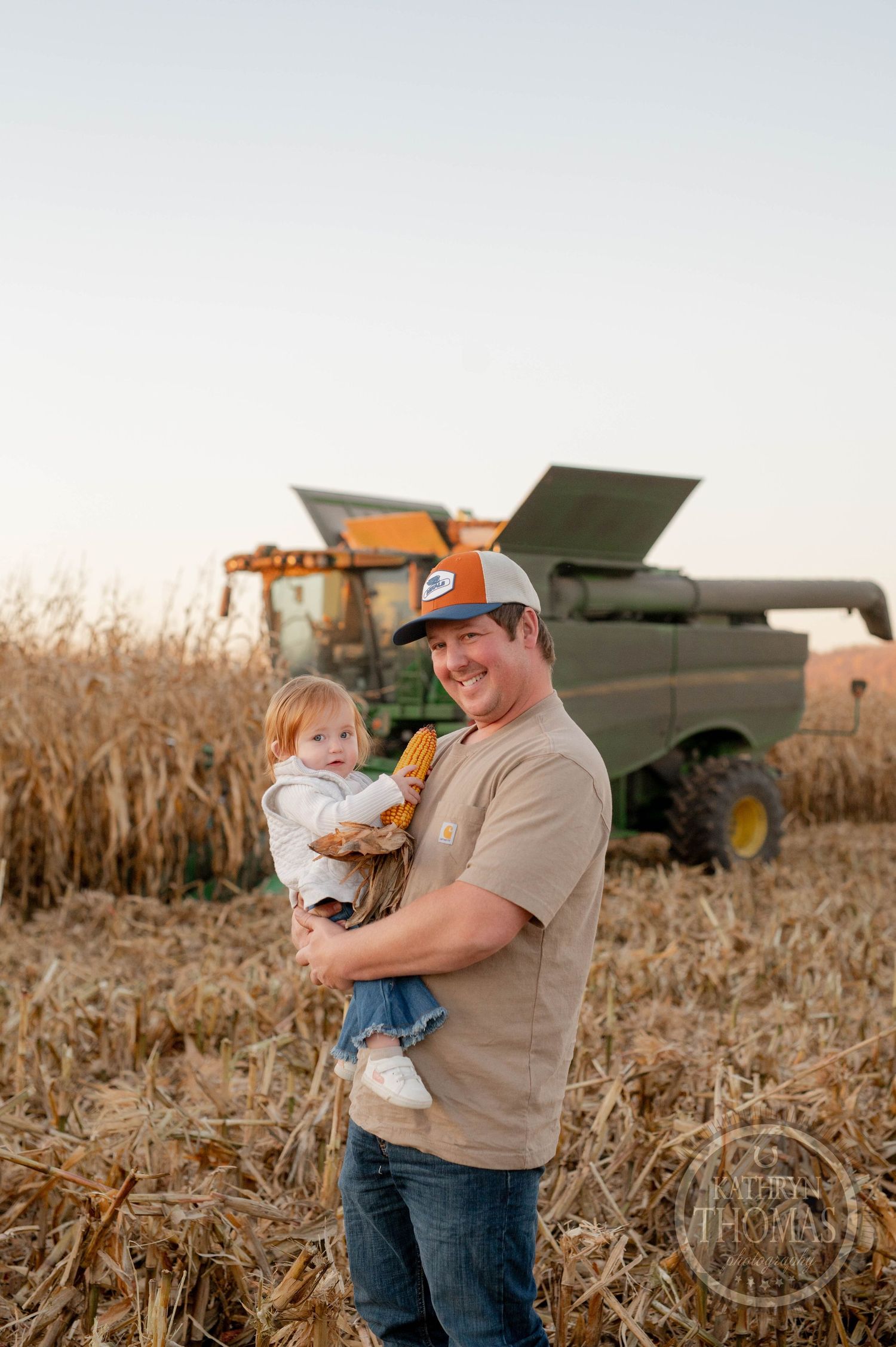 Farmer holds child against backdrop of combine harvester in cornfield at sunset.