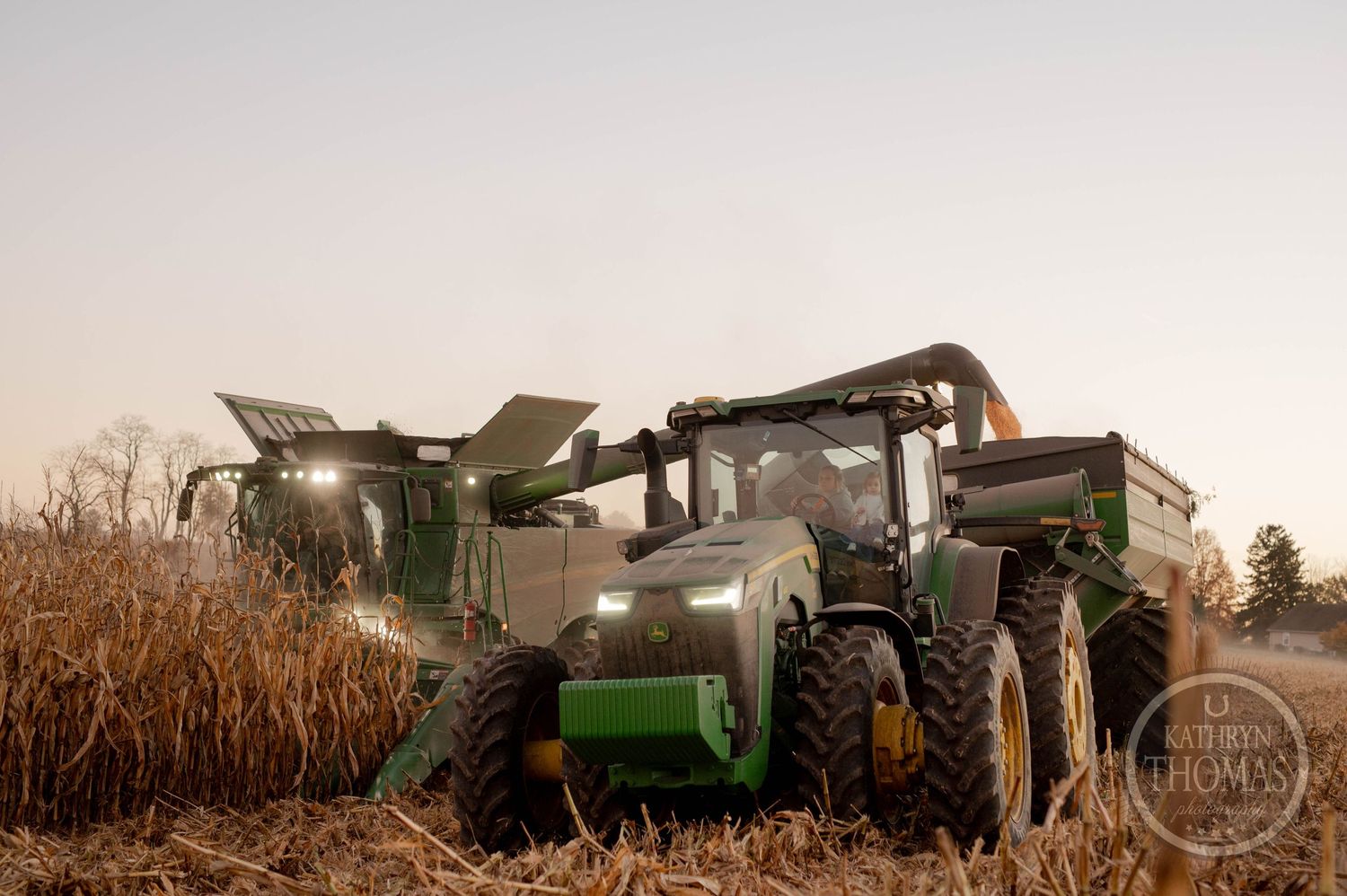 Modern combine harvester and tractor with grain cart harvesting corn at dusk.