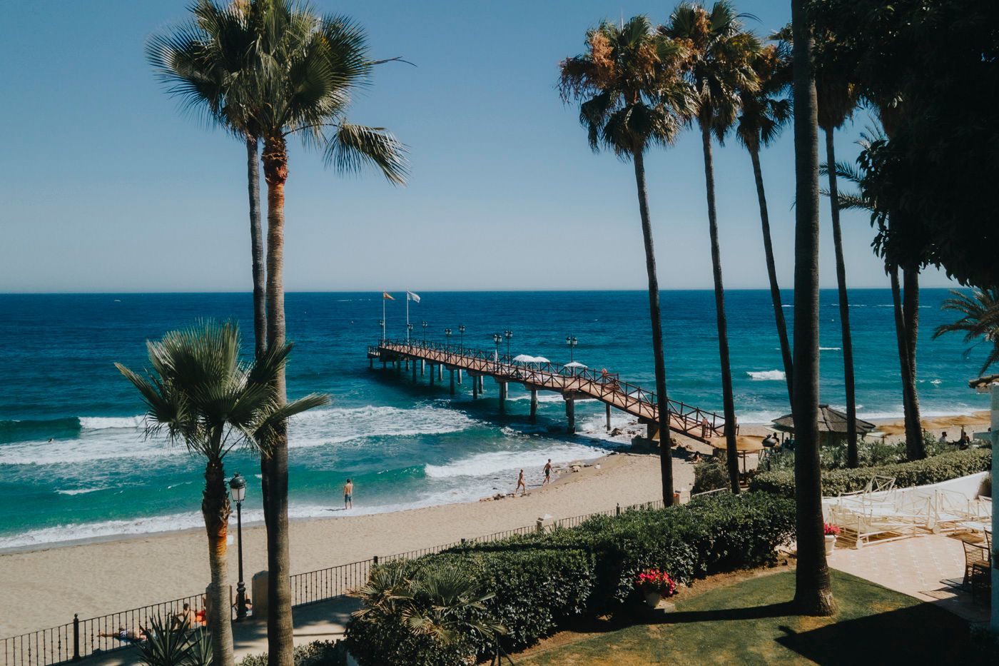 A scenic beach pier extends into turquoise ocean waters, framed by tall palm trees and white sandy shoreline in California.