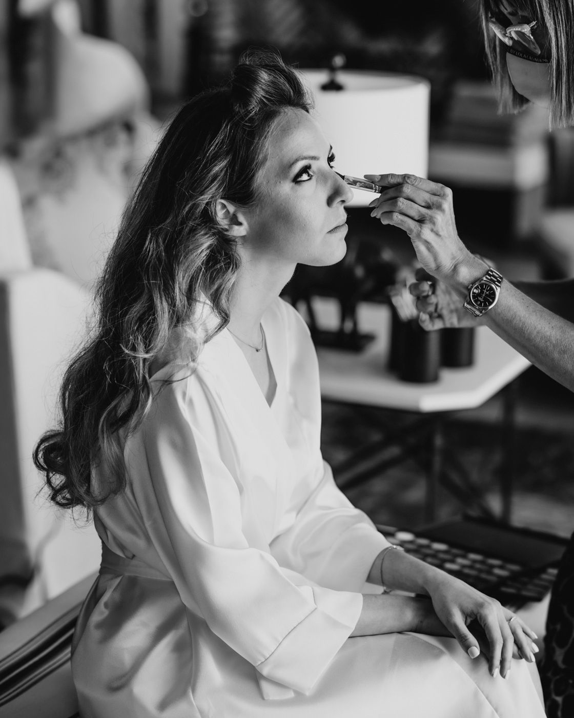 Black and white portrait showing someone getting makeup applied while wearing a white robe in a salon setting.