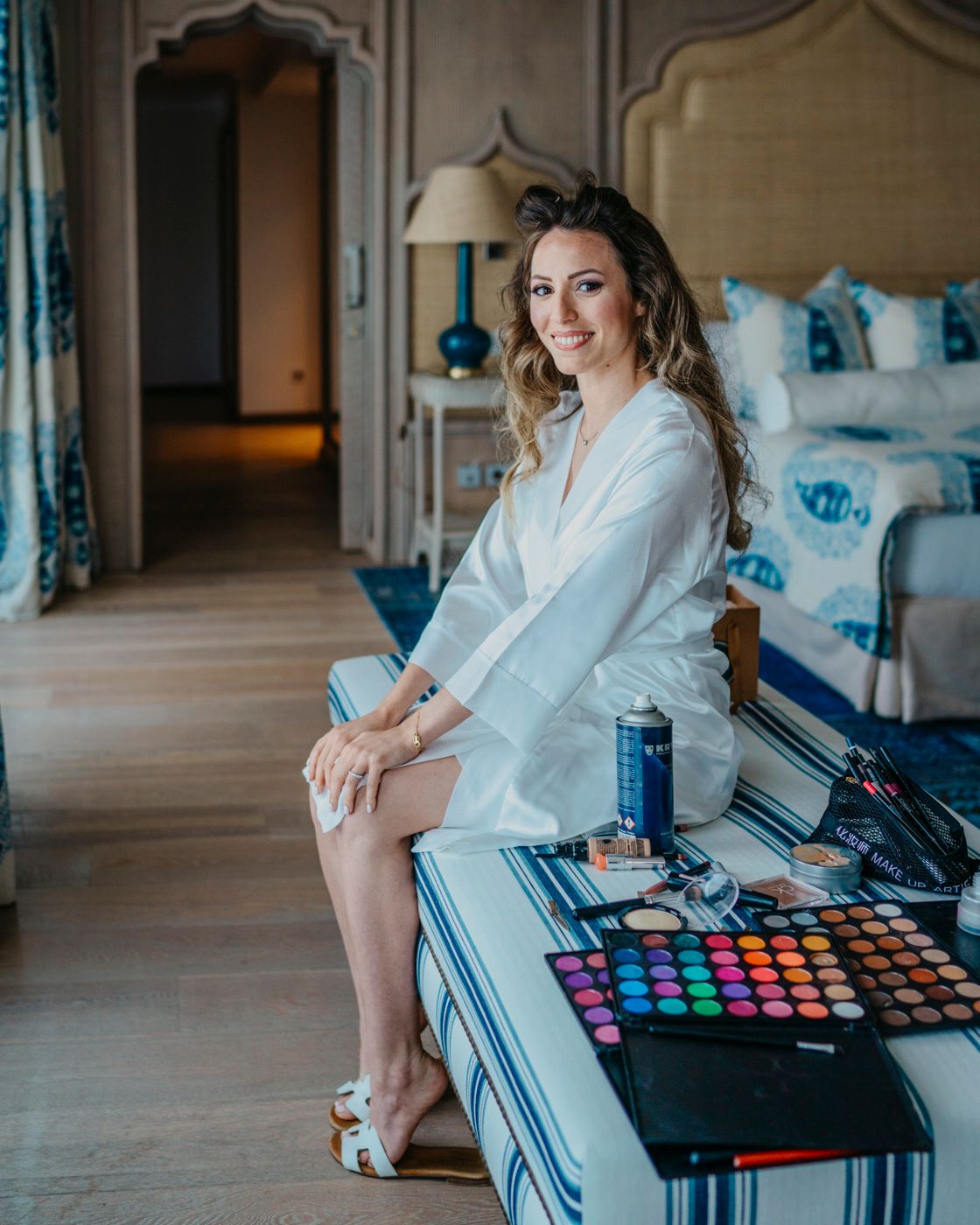 Professional makeup artist sits on edge of desk with cosmetics and colorful eyeshadow palette during wedding preparation.