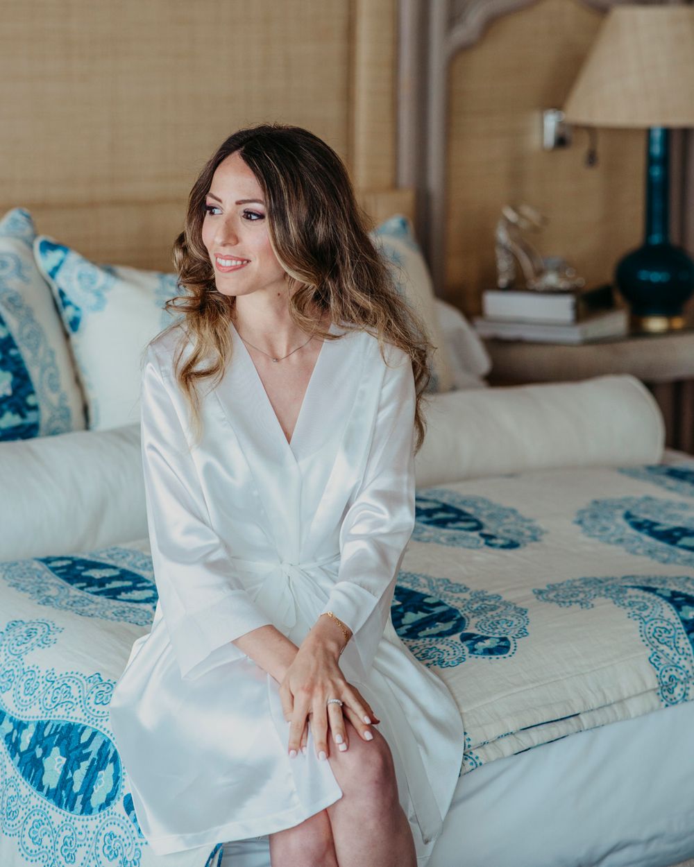 A person in a white silk robe sits on a bed with blue and white patterned bedding in a sunlit bedroom.