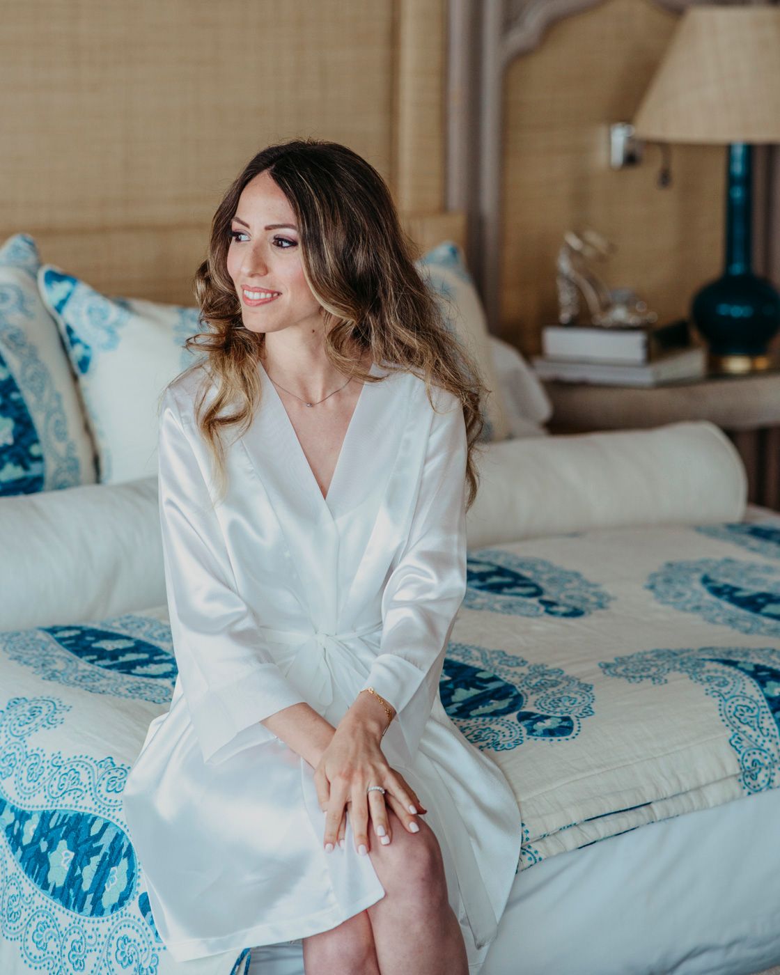 A person in a white silk robe sits on a bed with blue and white patterned bedding in a sunlit bedroom.