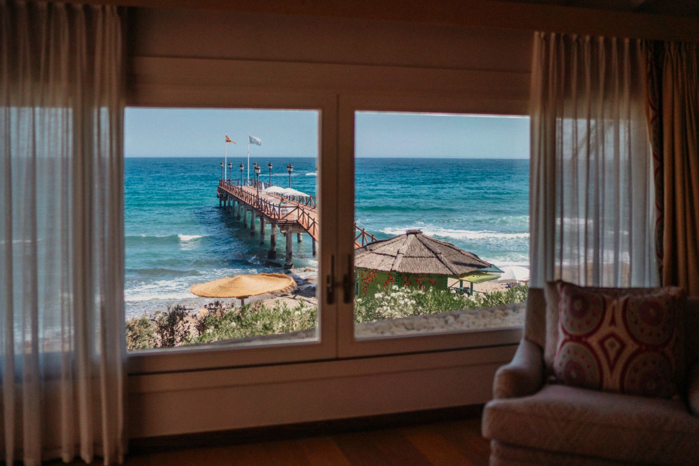 View of a wooden pier stretching into turquoise ocean waters, framed by curtained windows from inside a hotel room.