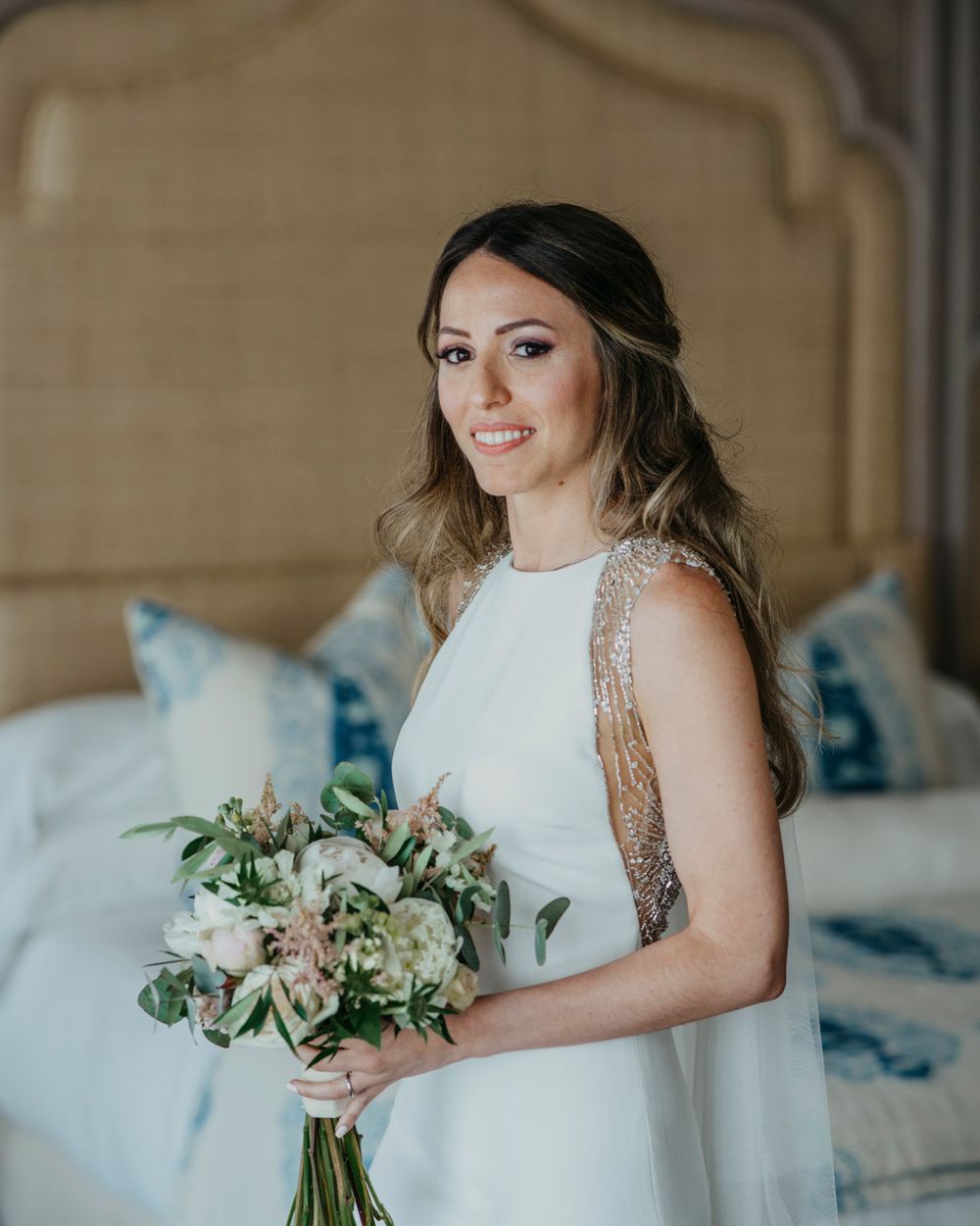 A bride in a sleeveless white dress holds a delicate bouquet of pale flowers against a soft beige bedroom backdrop.