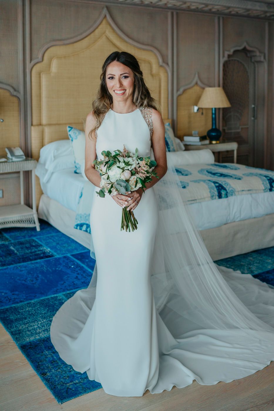A bride in an elegant white fitted wedding dress with a flowing train holds a green and white bouquet in a blue and white bedroom.