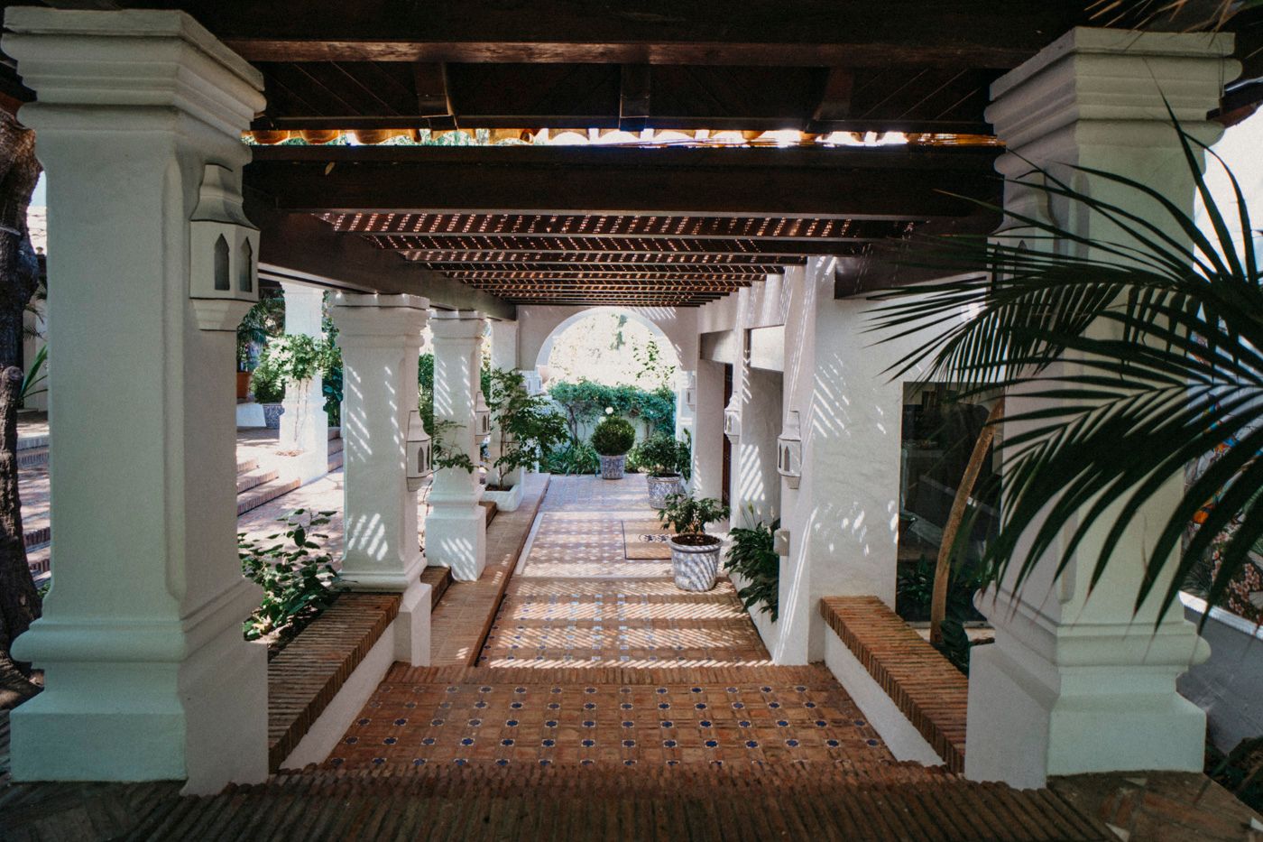 A covered walkway with white columns and terracotta tiles extends through a tropical garden setting.