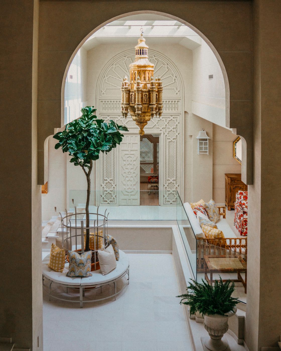 Elegant entryway with arched doorway, ornate gold chandelier and potted ficus tree on a curved white bench.