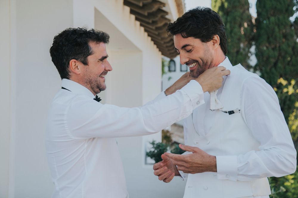 Two people in white dress shirts sharing a heartfelt moment and smiling while standing outdoors near a white wall.
