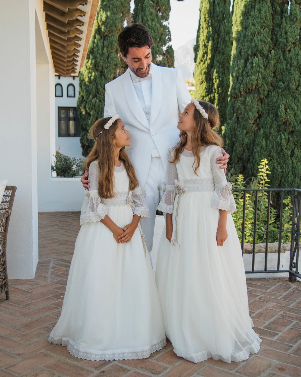 A person in a white suit stands with two small figures wearing ethereal white dresses and veils on a terrace with cypress trees.