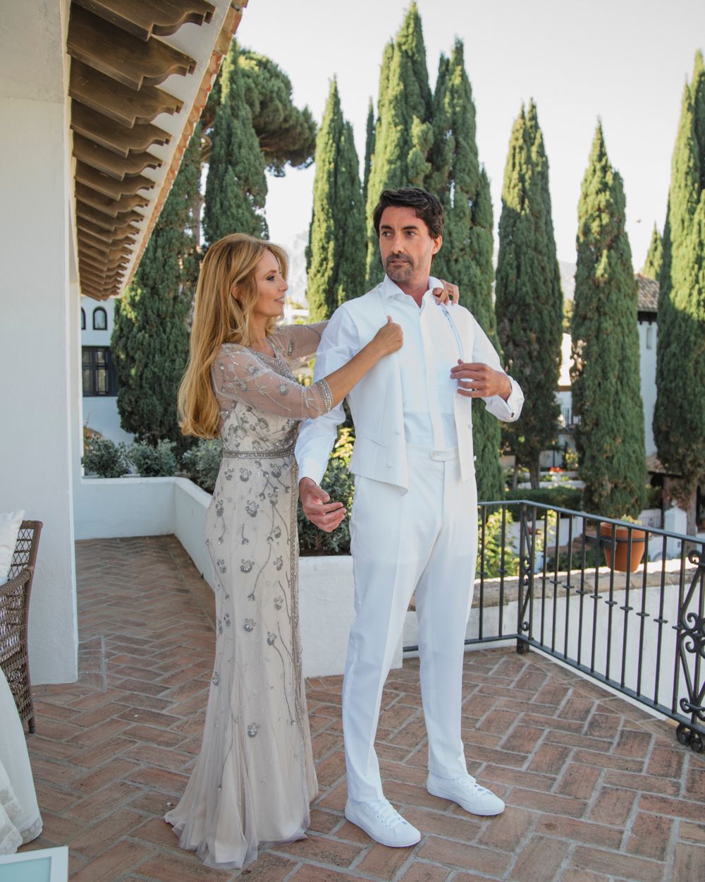A couple in elegant attire dance together on a Mediterranean terrace with tall cypress trees in the background.