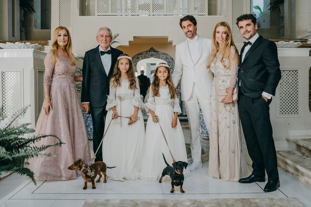 Elegant formal group portrait on white steps with two small dogs, dressed in white and pastel gowns and suits.