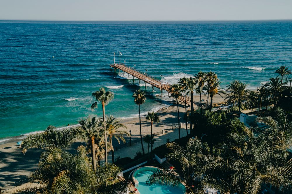 Aerial view of a coastal resort with palm trees, swimming pool and long pier extending into turquoise ocean waters.
