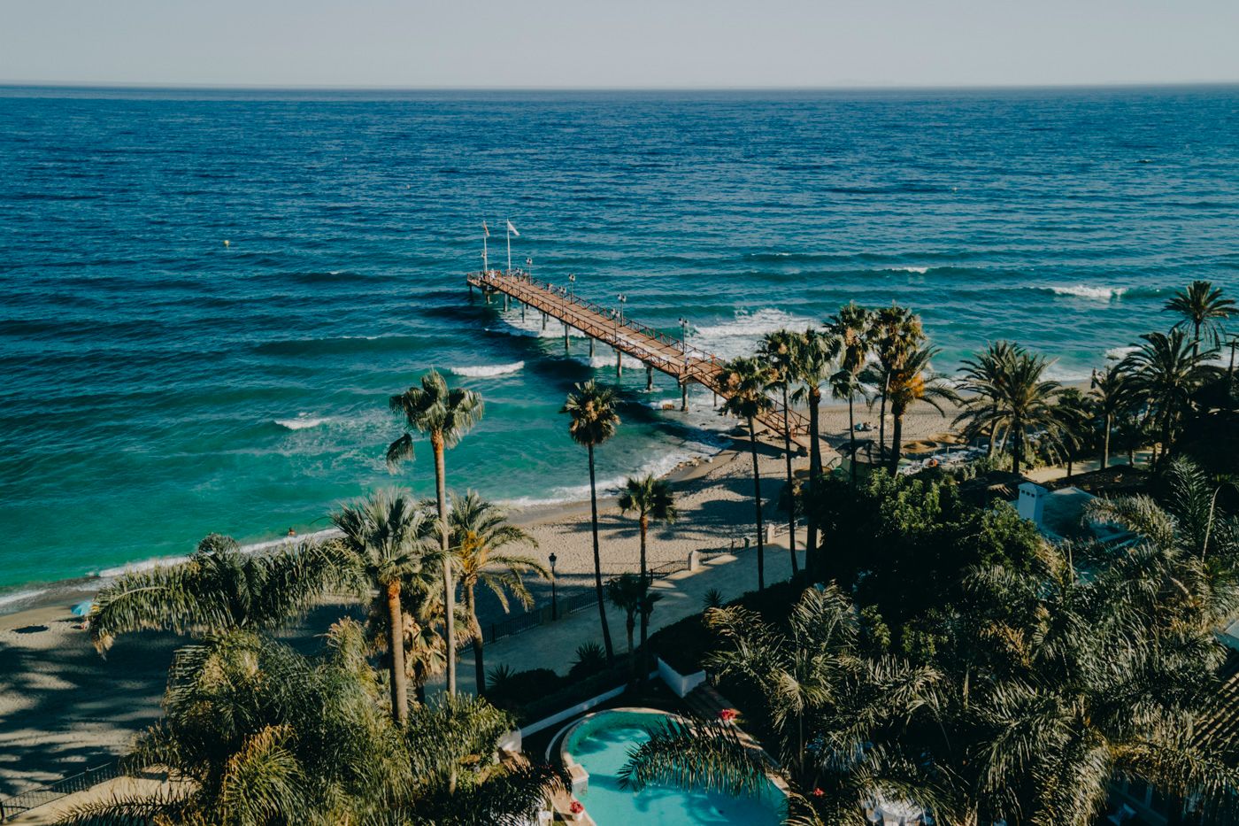 Aerial view of a coastal resort with palm trees, swimming pool and long pier extending into turquoise ocean waters.