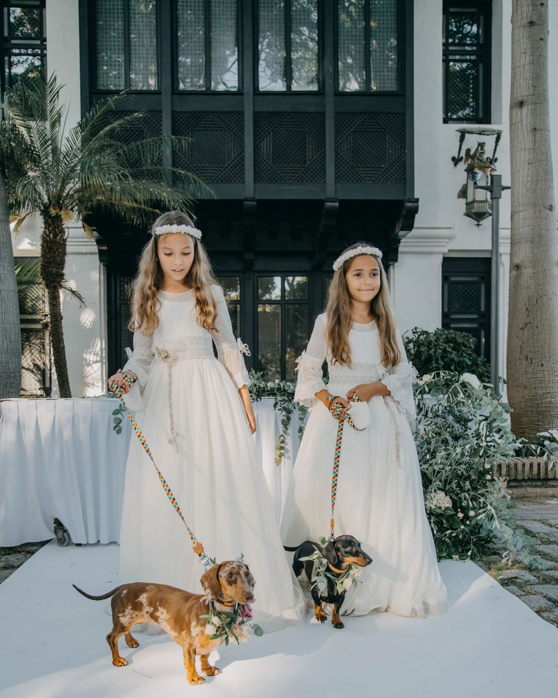 Two young attendants in white dresses stand with dachshund dogs on leashes outside an ornate building entrance.