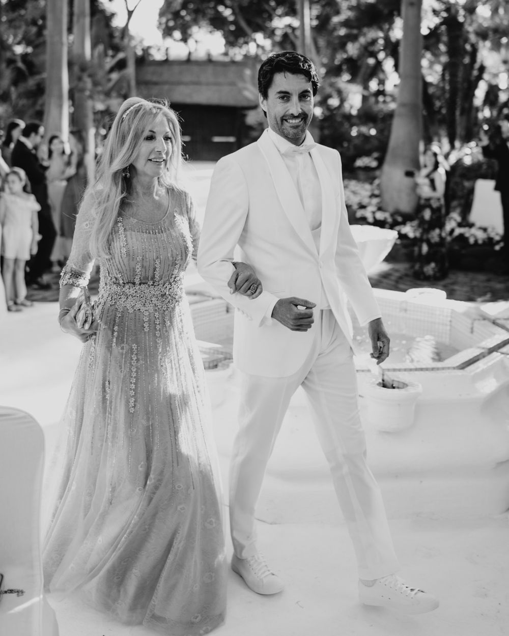 Black and white wedding photo showing a couple in white attire walking together at an outdoor ceremony.