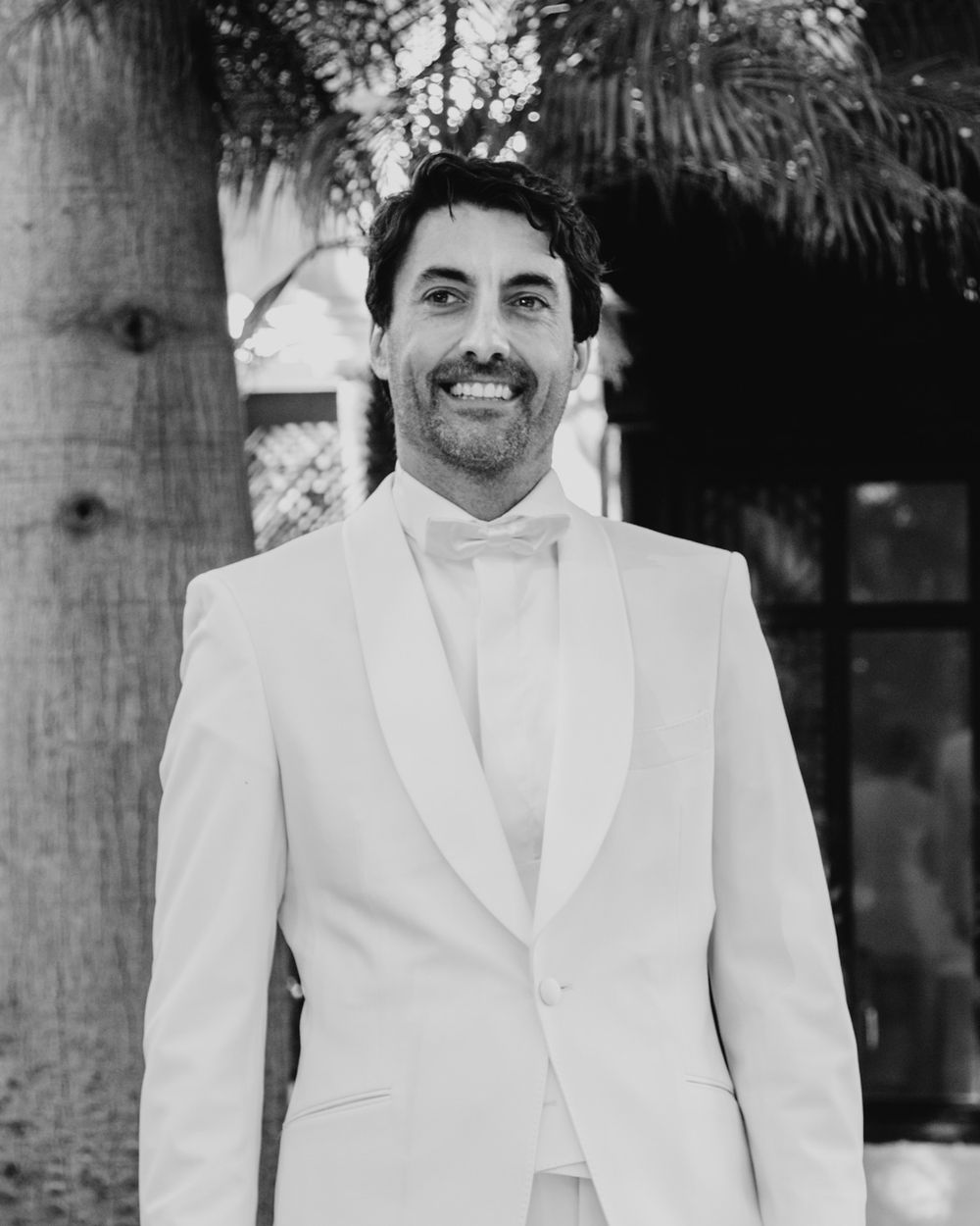 A groom smiling at a wedding wearing a crisp white tuxedo with a white bow tie against a palm tree background.