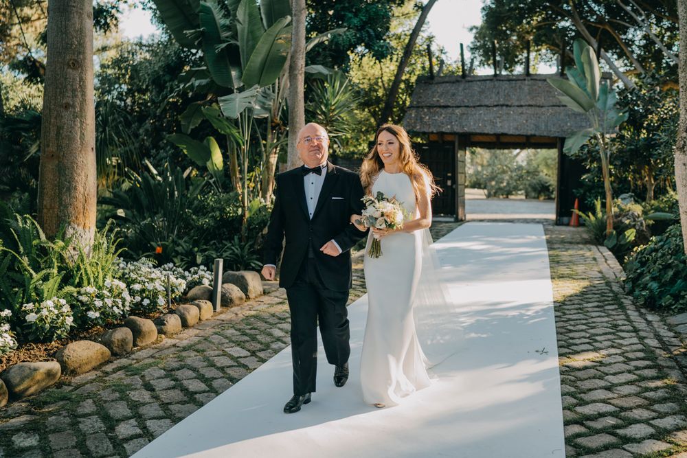 A couple walks down a tropical garden pathway with a wooden archway ahead, surrounded by lush greenery and palm trees.