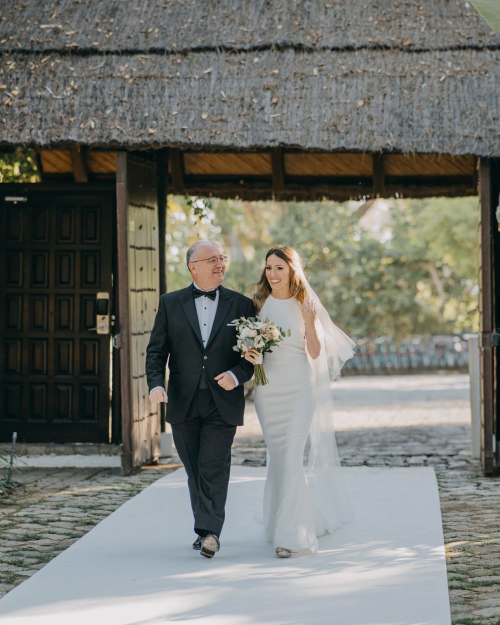 A bride in white gown walks down an outdoor aisle with a companion in formal black suit under a rustic wooden archway.