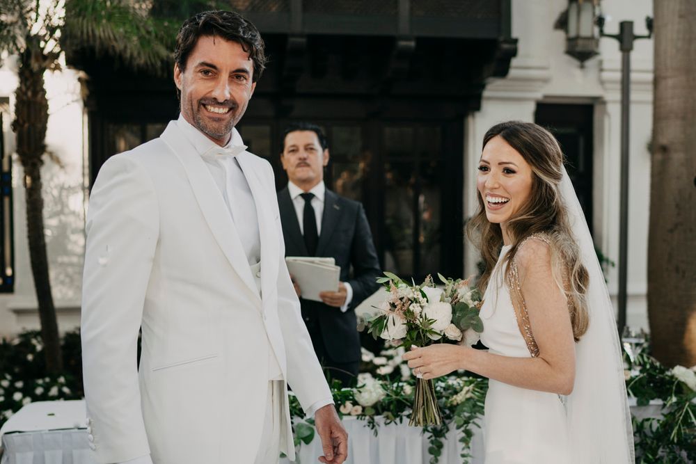 A wedding ceremony takes place outdoors with a couple in white attire and an officiant standing between ornate pillars.