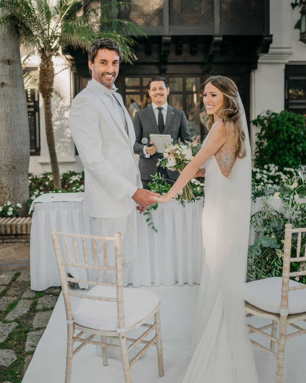 A couple exchanges vows during an intimate outdoor wedding ceremony with white chiavari chairs and tropical greenery.