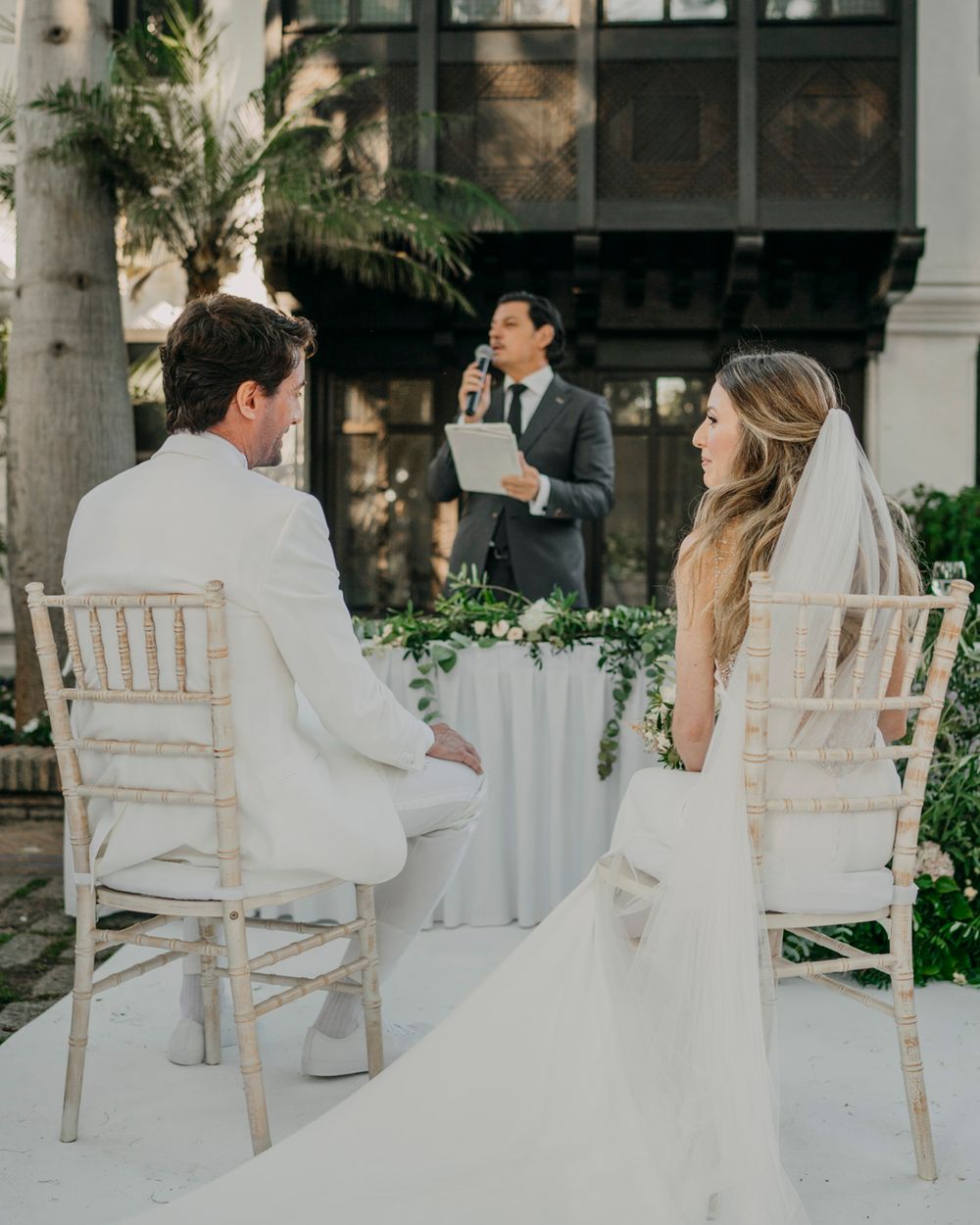 Couple sits in elegant white chairs at outdoor wedding ceremony while officiant delivers marriage vows on sunny day.