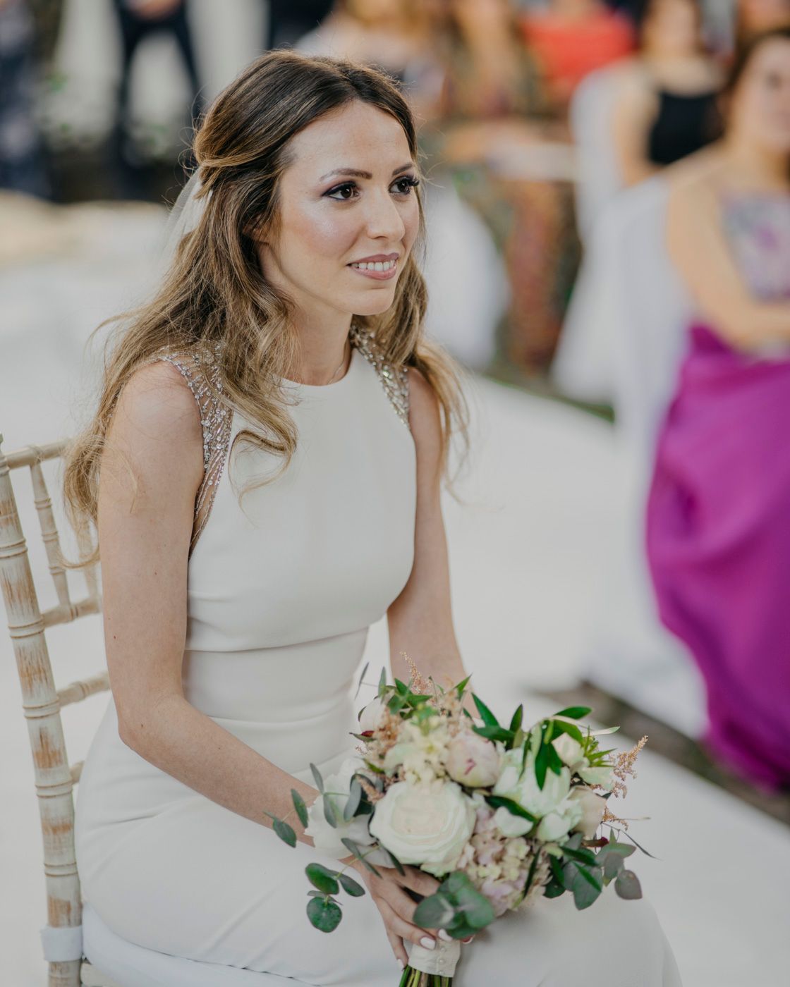 A bride in a sleek white dress sits holding a blush and cream bouquet with eucalyptus at a wedding celebration.
