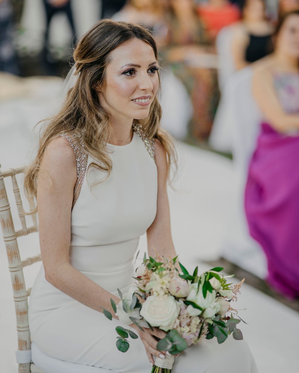 A bride in a sleek white dress sits holding a blush and cream bouquet with eucalyptus at a wedding celebration.