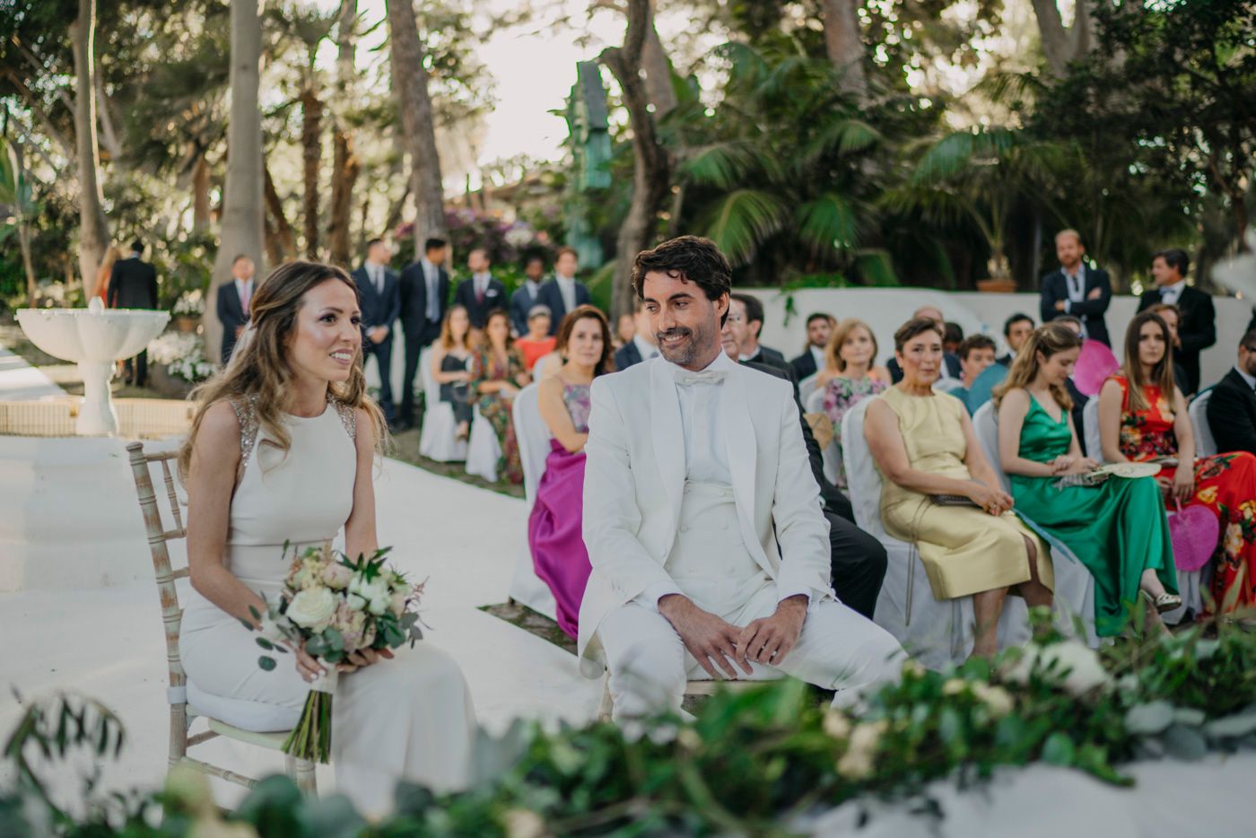 Wedding ceremony outdoors with couple seated at decorated table while guests in colorful attire look on.