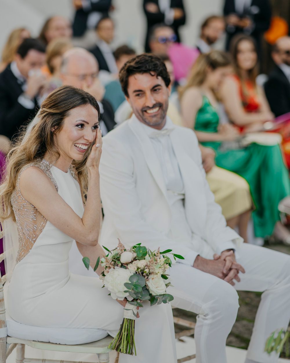 A couple at a wedding ceremony sits outdoors in all-white attire with a delicate white and green bridal bouquet.