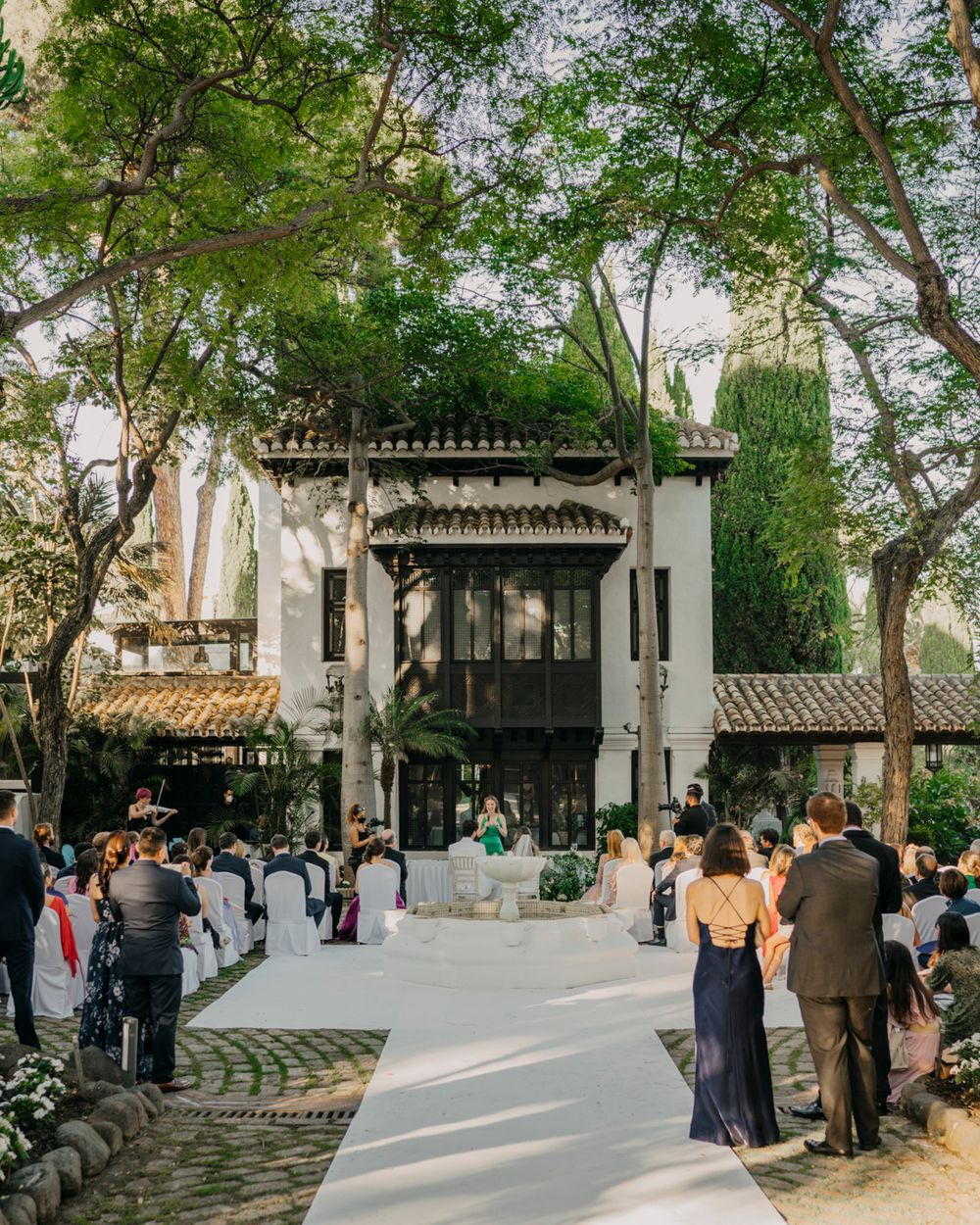 Elegant outdoor wedding ceremony setup with white chairs and grand gazebo framed by lush trees in a garden setting.