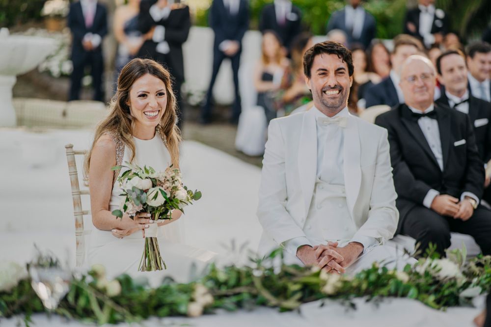 A bride in a white dress holds a bouquet while seated next to her groom in a light colored suit during an outdoor ceremony.