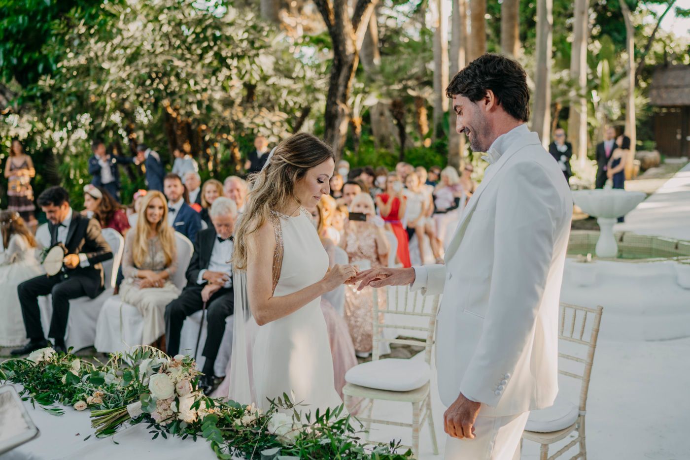 Outdoor garden wedding ceremony with floral table decorations and guests seated on white chairs in a tropical setting.