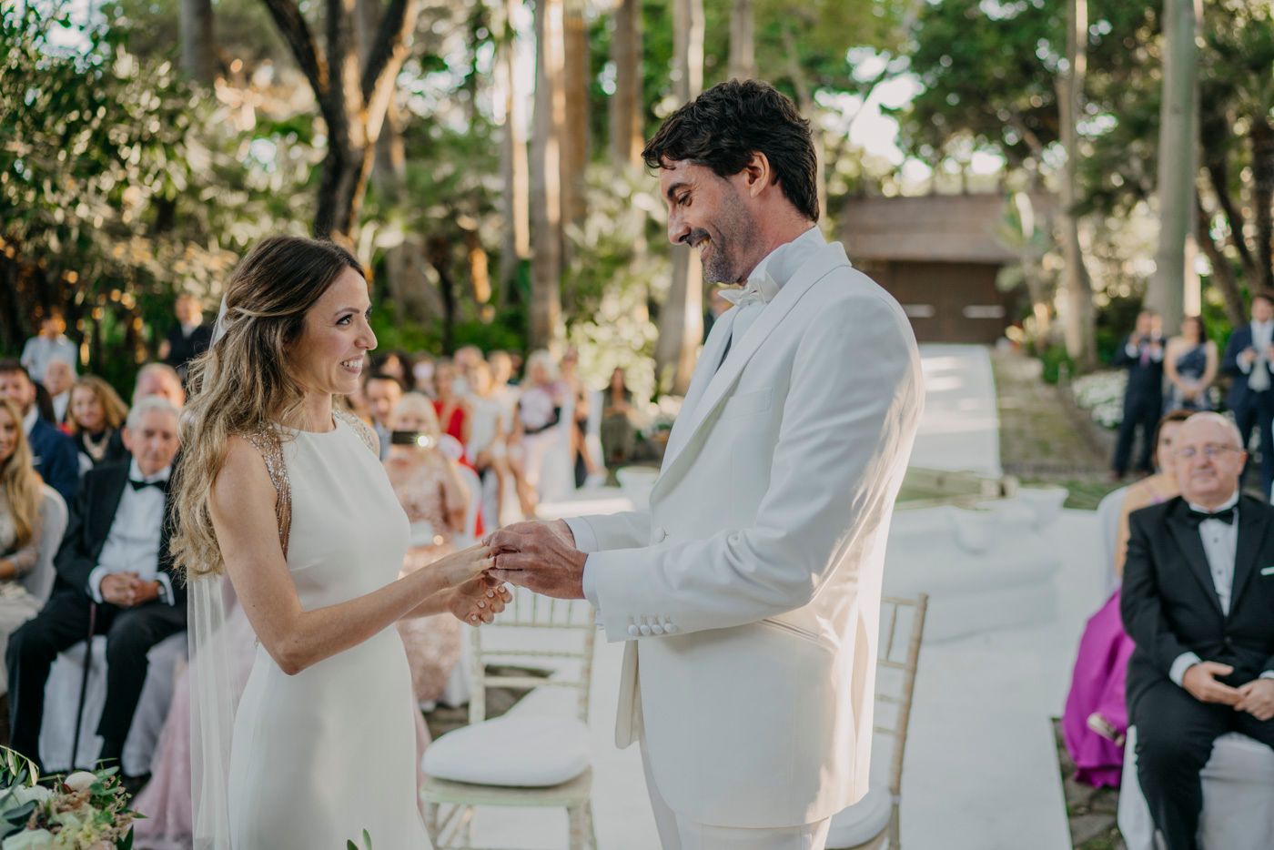 A romantic outdoor wedding ceremony with a couple exchanging rings in front of gathered guests in a garden setting.
