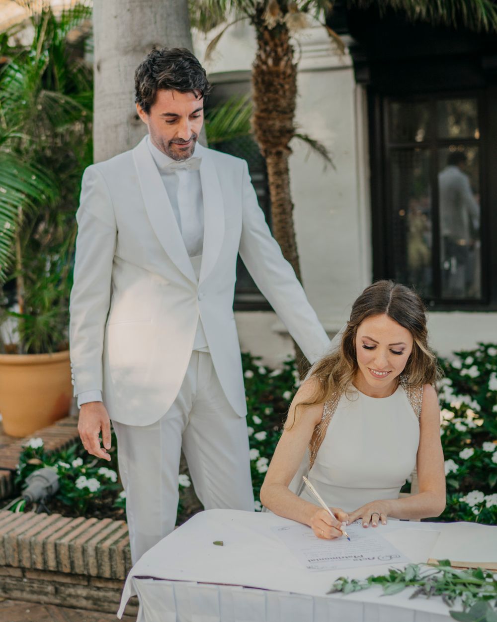 Two people in light-colored formal attire sign documents at an outdoor table surrounded by tropical landscaping.