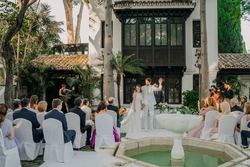 An outdoor wedding ceremony takes place at an elegant Spanish-style venue with white chairs arranged around a fountain courtyard.